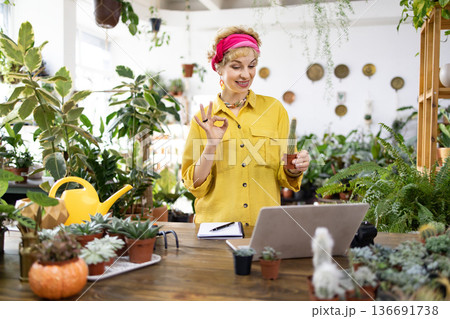 A woman in a yellow shirt holds a small cactus while gesturing during a video call in a plant shop A woman in a yellow shirt holds a small cactus while gesturing during a video call in a plant shop 136691738