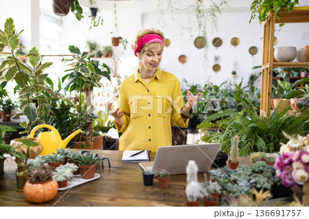 A woman in a yellow shirt gestures while speaking in a plant shop, surrounded by lush greenery and a laptop A woman in a yellow shirt gestures while speaking in a plant shop, surrounded by lush greenery and a laptop 136691757