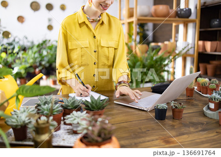 A woman in a yellow shirt works on a laptop and takes notes surrounded by plants and succulents A woman in a yellow shirt works on a laptop and takes notes surrounded by plants and succulents 136691764