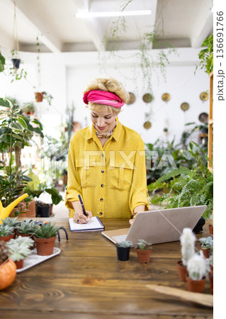 A woman in a yellow shirt works on a laptop and takes notes in a plant shop surrounded by greenery A woman in a yellow shirt works on a laptop and takes notes in a plant shop surrounded by greenery 136691766