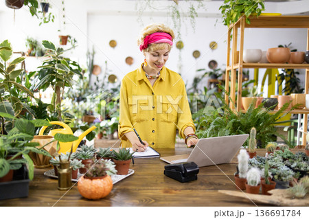 A woman in a yellow shirt works on a laptop surrounded by lush plants in a bright shop A woman in a yellow shirt works on a laptop surrounded by lush plants in a bright shop 136691784