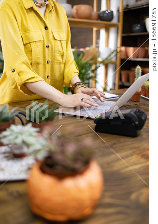 A woman in a yellow shirt works on a laptop in a plant shop, surrounded by succulents and other plants 136691785
