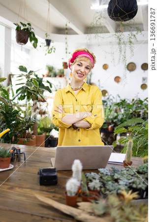 A smiling woman stands in her plant shop with her arms crossed, surrounded by greenery A smiling woman stands in her plant shop with her arms crossed, surrounded by greenery 136691787