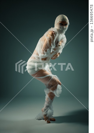 Conceptual portrait of a person wrapped like a mummy in white bandages, struggling in a constrained pose. Conceptual portrait of a person wrapped like a mummy in white bandages, struggling in a constrained pose. 136695478