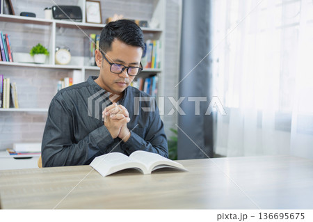 Young man in glasses praying with hands clasped in front of an open book at a wooden table in a bright indoor space with natural light and shelves Young man in glasses praying with hands clasped in front of an open book at a wooden table in a bright indoor space with natural light and shelves 136695675
