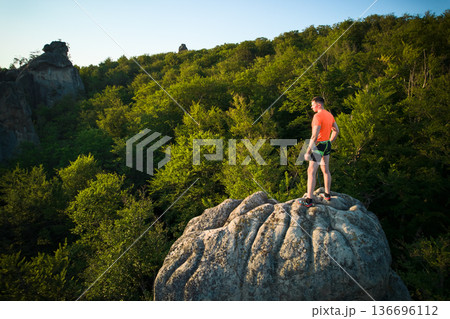Aerial view of climber standing atop rock formation at Dovbush Rocks in Carpathian mountains, Ukraine. Sun sets, casting warm glow over lush, green forest, distant hills, creating breathtaking view. Aerial view of climber standing atop rock formation at Dovbush Rocks in Carpathian mountains, Ukraine. Sun sets, casting warm glow over lush, green forest, distant hills, creating breathtaking view. 136696112