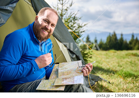 Portrait of smiling man sits in tourist tent's entrance. Tourist holds compass and contemplates his route, surrounded by lush green field with trees and mountains in distance under cloudy sky. 136696116