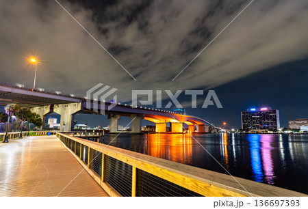 The Acosta Bridge spans the St. Johns River in Jacksonville, Florida. The illuminated bridge and city skyline are viewed from the wooden boardwalk of the Northbank Riverwalk at night. 136697393