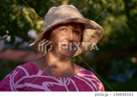 a senior woman in pink dress and straw hat enjoying golden hour sunset in the garden 136698101