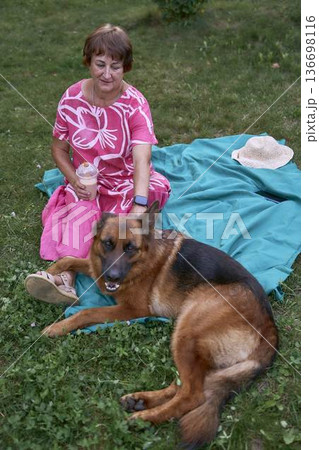 a Senior woman relaxing on a picnic blanket with german shepherd dog and coffee in a garden a Senior woman relaxing on a picnic blanket with german shepherd dog and coffee in a garden 136698116