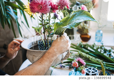 Man arranges pink and purple asters in vintage white urn using wire mesh. dopamine decor flowers, bright floral energy, mood boosting bouquets, colorful flower therapy Man arranges pink and purple asters in vintage white urn using wire mesh. dopamine decor flowers, bright floral energy, mood boosting bouquets, colorful flower therapy 136699548