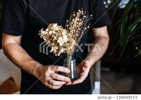 Person holds small glass vase with dried hydrangea and stalks against black shirt. decaying floral beauty, dried flower aesthetics, beauty in imperfection flowers Person holds small glass vase with dried hydrangea and stalks against black shirt. decaying floral beauty, dried flower aesthetics, beauty in imperfection flowers 136699555