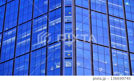 Big glowing windows in modern office building at night, in rows of windows light shines. Fragment of the glass facade of a modern corporate building at night. Modern glass office in city. Big glowing windows in modern office building at night, in rows of windows light shines. Fragment of the glass facade of a modern corporate building at night. Modern glass office in city. 136699740
