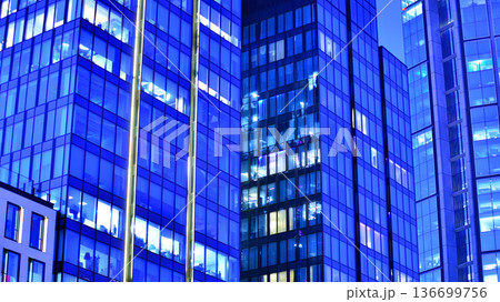 Big glowing windows in modern office building at night, in rows of windows light shines. Fragment of the glass facade of a modern corporate building at night. Modern glass office  in city. 136699756