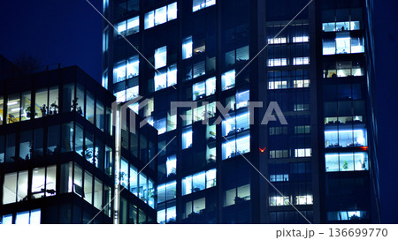 Big glowing windows in modern office building at night, in rows of windows light shines. Fragment of the glass facade of a modern corporate building at night. Modern glass office  in city. 136699770