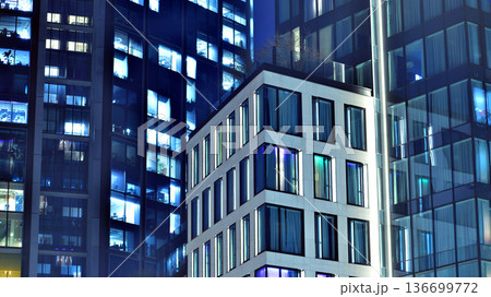 Big glowing windows in modern office building at night, in rows of windows light shines. Fragment of the glass facade of a modern corporate building at night. Modern glass office in city. Big glowing windows in modern office building at night, in rows of windows light shines. Fragment of the glass facade of a modern corporate building at night. Modern glass office in city. 136699772