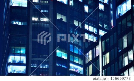 Big glowing windows in modern office building at night, in rows of windows light shines. Fragment of the glass facade of a modern corporate building at night. Modern glass office in city. Big glowing windows in modern office building at night, in rows of windows light shines. Fragment of the glass facade of a modern corporate building at night. Modern glass office in city. 136699778
