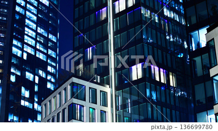 Big glowing windows in modern office building at night, in rows of windows light shines. Fragment of the glass facade of a modern corporate building at night. Modern glass office in city. Big glowing windows in modern office building at night, in rows of windows light shines. Fragment of the glass facade of a modern corporate building at night. Modern glass office in city. 136699780