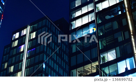 Big glowing windows in modern office building at night, in rows of windows light shines. Fragment of the glass facade of a modern corporate building at night. Modern glass office in city. Big glowing windows in modern office building at night, in rows of windows light shines. Fragment of the glass facade of a modern corporate building at night. Modern glass office in city. 136699782