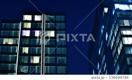 Big glowing windows in modern office building at night, in rows of windows light shines. Fragment of the glass facade of a modern corporate building at night. Modern glass office in city. Big glowing windows in modern office building at night, in rows of windows light shines. Fragment of the glass facade of a modern corporate building at night. Modern glass office in city. 136699786