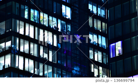 Big glowing windows in modern office building at night, in rows of windows light shines. Fragment of the glass facade of a modern corporate building at night. Modern glass office in city. Big glowing windows in modern office building at night, in rows of windows light shines. Fragment of the glass facade of a modern corporate building at night. Modern glass office in city. 136699794