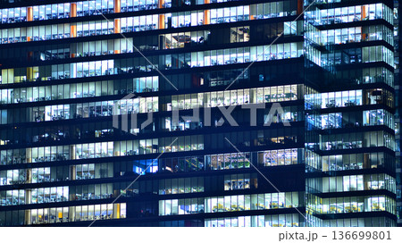 Big glowing windows in modern office building at night, in rows of windows light shines. Fragment of the glass facade of a modern corporate building at night. Modern glass office in city. Big glowing windows in modern office building at night, in rows of windows light shines. Fragment of the glass facade of a modern corporate building at night. Modern glass office in city. 136699801