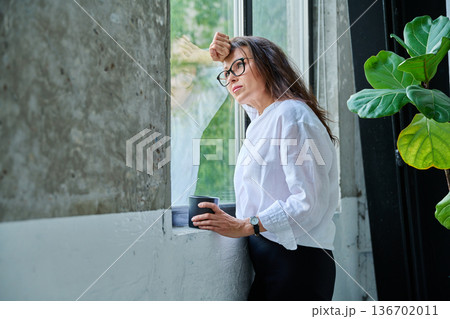 Middle-aged serious woman with cup looking at window 136702011