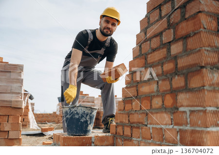 In uniform and hard hat. Construction worker is at job with bricks outdoors In uniform and hard hat. Construction worker is at job with bricks outdoors 136704072