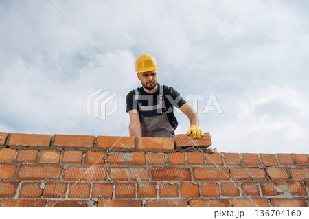 Against cloudy sky, making wall. Construction worker is at job with bricks outdoors Against cloudy sky, making wall. Construction worker is at job with bricks outdoors 136704160