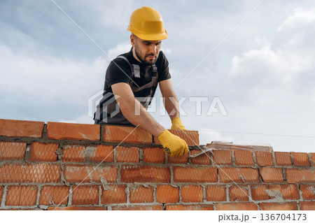 Against cloudy sky, making wall. Construction worker is at job with bricks outdoors 136704173
