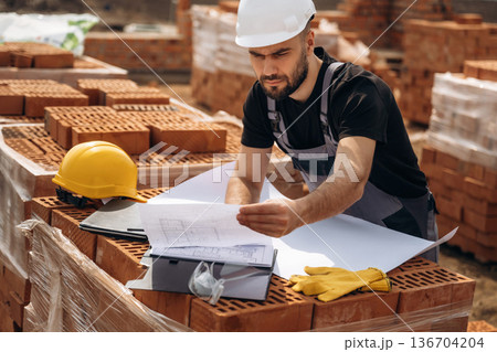 Project plan on the paper. Construction worker is at job with bricks outdoors Project plan on the paper. Construction worker is at job with bricks outdoors 136704204