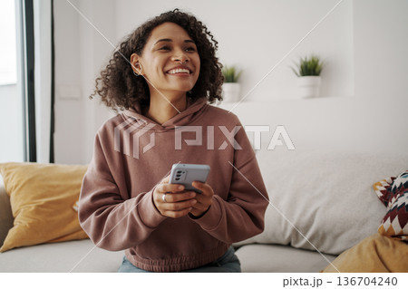 Woman With Bright Expression Engaging In Video Conversation Amid Plantfilled Room And Soft Cushions 136704240