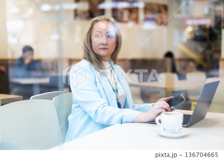 Business lunch - businesswoman sits at table in cafe by window with a laptop and drinks coffee. View through window 136704665
