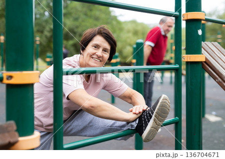 elderly woman stretches on the bars in an outdoor sports area elderly woman stretches on the bars in an outdoor sports area 136704671