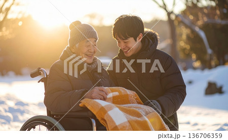 冬の公園で車椅子の高齢者に膝掛けをかけ笑顔で寄り添う若い男性介護士 136706359