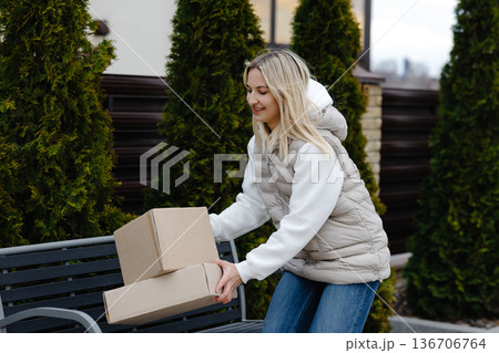 Woman holding cardboard boxes standing next to a bench in front of her house 136706764