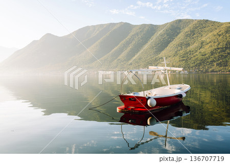 A serene red boat peacefully floating on calm waters surrounded by mountains in early morning light 136707719