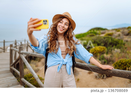 Young woman with curly hair wears a stylish hat and waves while taking a selfie. Travel, tourism 136708089