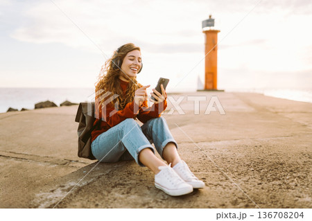 Happy woman listening nice music by waterfront while wearing headphones. Travel, blogging concept. Happy woman listening nice music by waterfront while wearing headphones. Travel, blogging concept. 136708204