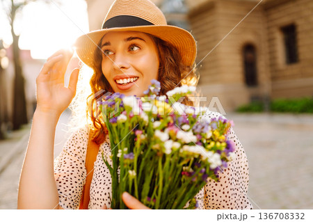 Beautiful young woman in summer style outfit smiling happy walking with flowers in city street 136708332