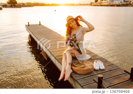 Young woman in dress sitting on wooden pier near pond. Rustic style of wedding. Travel, tourism Young woman in dress sitting on wooden pier near pond. Rustic style of wedding. Travel, tourism 136708408