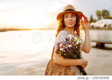 Happy woman in dress sitting on wooden pier near pond and clutched bouquet. Rustic style of wedding. 136708409
