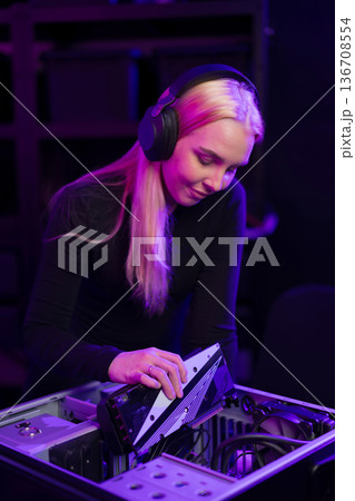 Female gamer builds gaming PC under purple lights with headphones on and focused expression 136708554