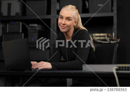 Confident female IT technician at a desk with a laptop and network gear in a technical office 136708563