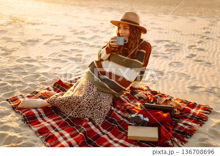 Cute woman sits on the beach drinks a hot drink from a thermos. Picnic, weekend, relax and lifestyle 136708696