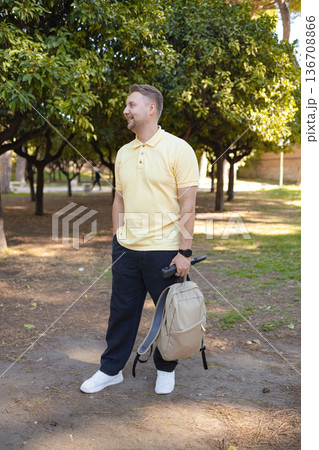 Man Outdoors Holding Backpack in Green Park on Sunny Day. Male tourist or city walker enjoying nature in an urban park. Summer daylight, trees and greenery, relaxed atmosphere. Man Outdoors Holding Backpack in Green Park on Sunny Day. Male tourist or city walker enjoying nature in an urban park. Summer daylight, trees and greenery, relaxed atmosphere. 136708866