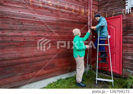 Man and woman over 50 years old are hanging garland on external wall of rural house. 136709172