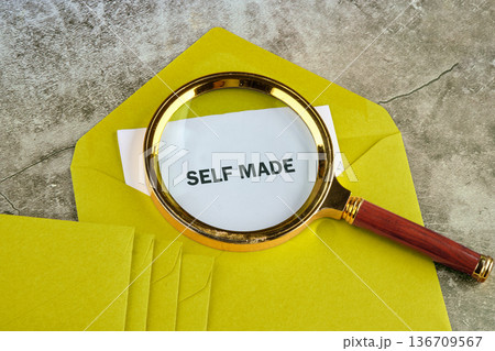A golden magnifying glass highlights self made on a white paper, nestled within a vibrant yellow envelope, resting upon a textured grey table A golden magnifying glass highlights self made on a white paper, nestled within a vibrant yellow envelope, resting upon a textured grey table 136709567