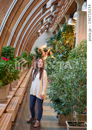 Woman in a white sweater looking up in greenhouse with various plants. Botanical garden interior design and environment Woman in a white sweater looking up in greenhouse with various plants. Botanical garden interior design and environment 136710234