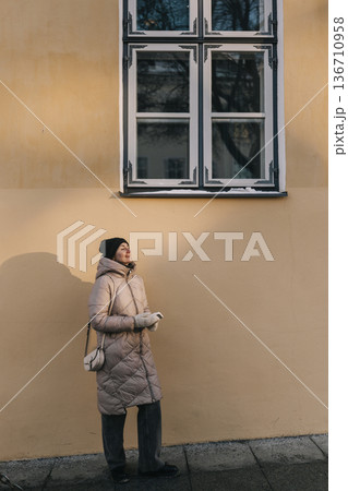 Woman stands by wall under window while looking upward in cool weather during daytime Woman stands by wall under window while looking upward in cool weather during daytime 136710958